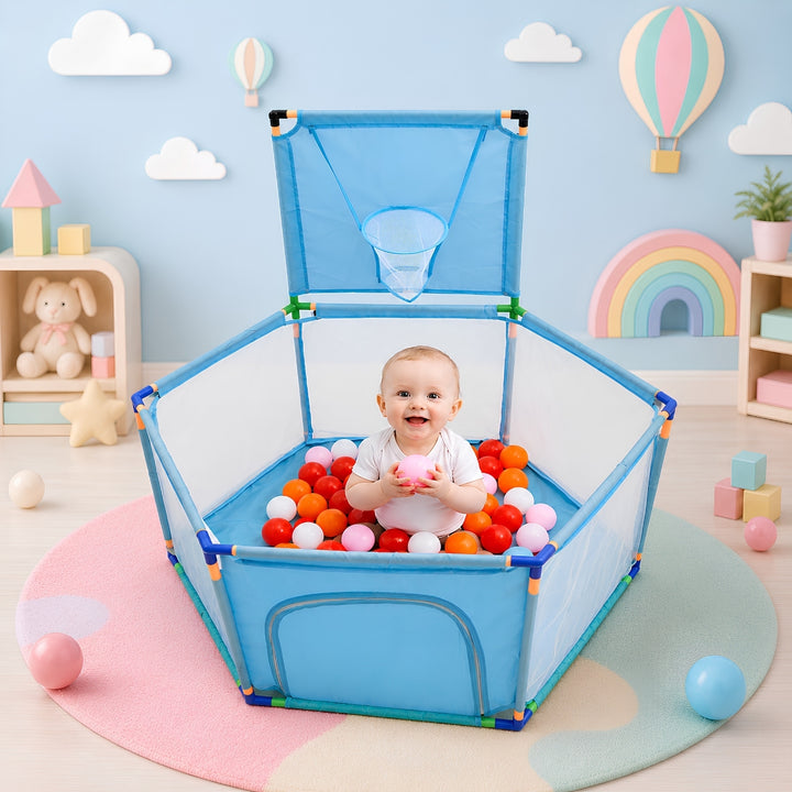 A baby sits inside a blue and white hexagonal ball pit playpen with an attached small basketball hoop, surrounded by colorful plastic balls in a pastel nursery room.