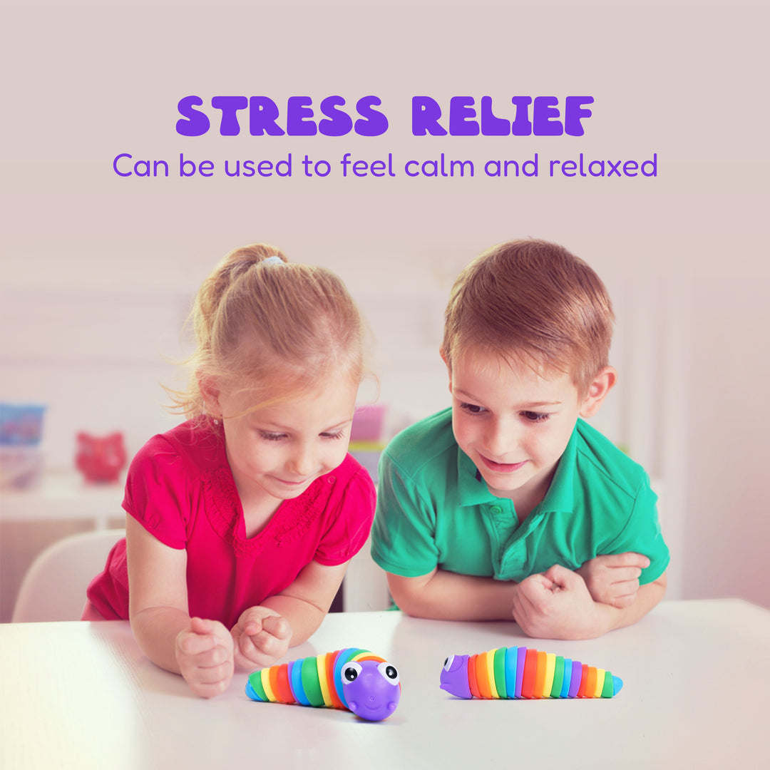 Two children, a girl in a pink shirt and a boy in a green shirt, looking at a colorful stress relief toy on a white table. The toy consists of two rainbow-striped caterpillar-like pieces with purple heads and black eyes.